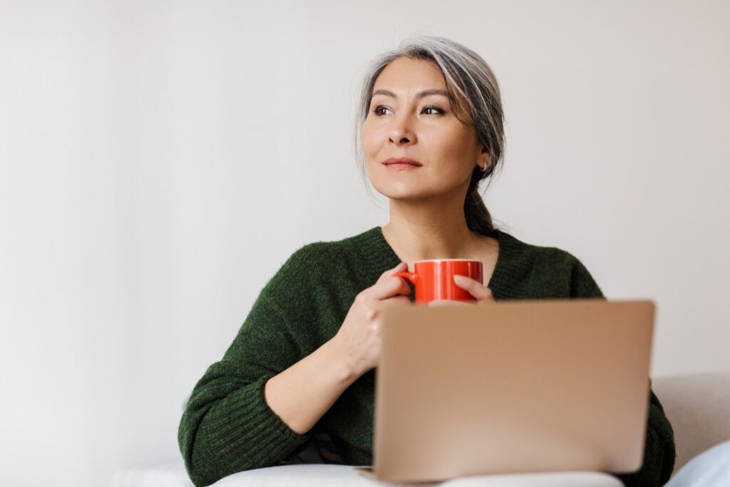 youthful looking middle aged woman holding mug while working on laptop